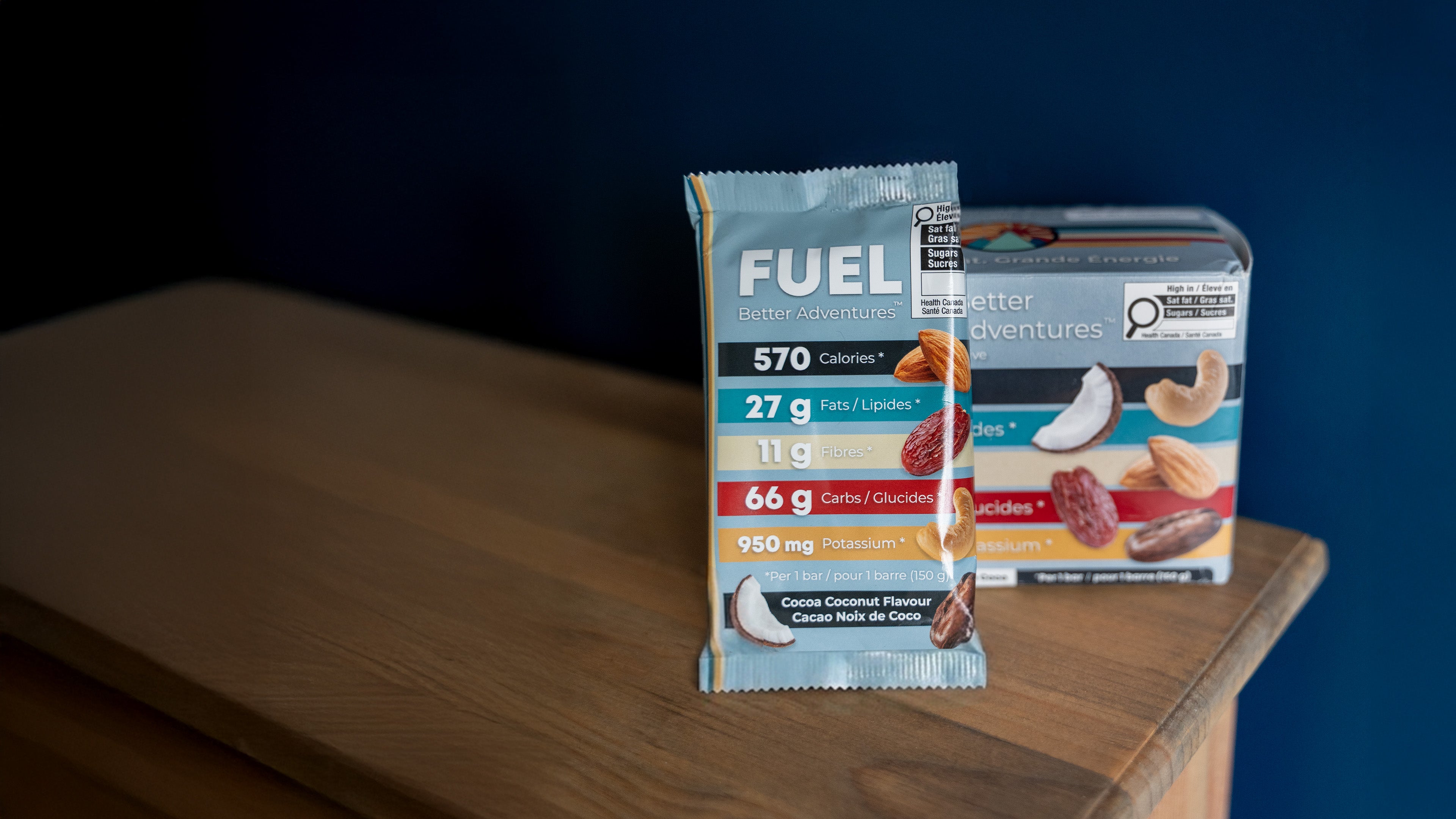 A single FUEL protein bar packet stands on a wooden table with its branded box behind it against a dark blue background.